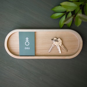 Può includere: Un vassoio di legno con un cartoncino grigio su cui è scritto "OKU" e un simbolo di albero stilizzato. Due chiavi sono sul vassoio.