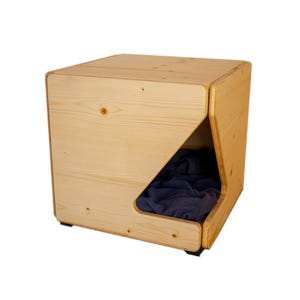 Pu&ograve; includere: Un lettino per cani in legno con una coperta blu all'interno. Il lettino ha la forma di un cubo con un'apertura triangolare su un lato.