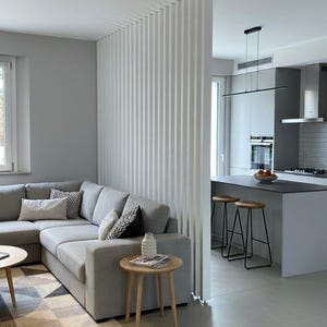 Peut inclure: Un salon avec un canap&eacute; d'angle gris, un tapis &agrave; motifs g&eacute;om&eacute;triques et une table d'appoint ronde en bois avec un petit vase blanc. Une cloison ajour&eacute;e &agrave; lattes verticales blanches s&eacute;pare le salon d'une cuisine avec &icirc;lot gris et tabourets.