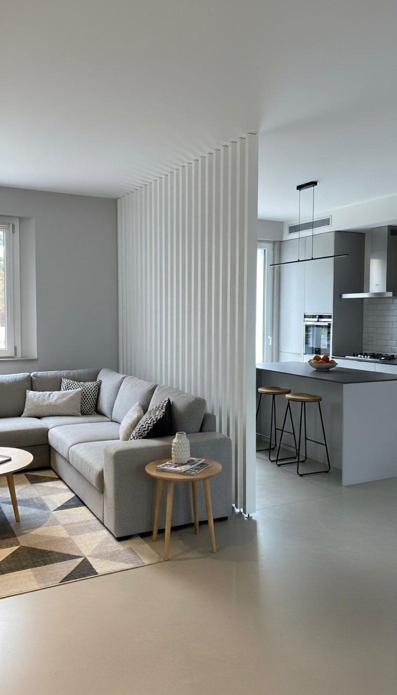 Peut inclure: Un salon avec un canap&eacute; d'angle gris, un tapis &agrave; motifs g&eacute;om&eacute;triques et une table d'appoint ronde en bois avec un petit vase blanc. Une cloison ajour&eacute;e &agrave; lattes verticales blanches s&eacute;pare le salon d'une cuisine avec &icirc;lot gris et tabourets.
