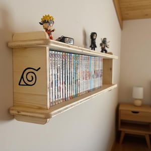 Può includere: Libreria a parete in legno con un simbolo a spirale nero e una collezione di manga. La mensola è decorata con statuette di personaggi anime. La mensola è in legno chiaro.