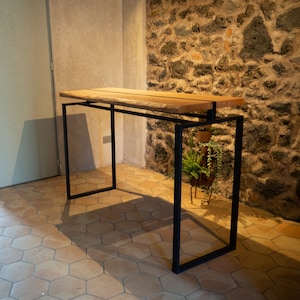 Puede incluir: Una mesa de madera moderna con borde natural, soportada por una estructura de metal negro. La encimera es de madera clara. La mesa está en una habitación con pared de piedra y suelo de baldosas.