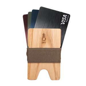 Op de afbeelding: Een houten kaarthouder met een bruin elastiek. De houder heeft een uitsparing voor kaarten en een logo met de tekst "OKU". De houder houdt een stapel kaarten vast, waaronder een zwarte VISA Platinum kaart.