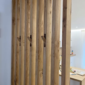 Peut inclure: Un s&eacute;parateur de pi&egrave;ce compos&eacute; de lattes de bois verticales brun clair dispos&eacute;es en parall&egrave;le. Plusieurs de ces lattes pr&eacute;sentent des crochets en bois inclin&eacute;s int&eacute;gr&eacute;s qui d&eacute;passent du centre, offrant une solution de rangement fonctionnelle.