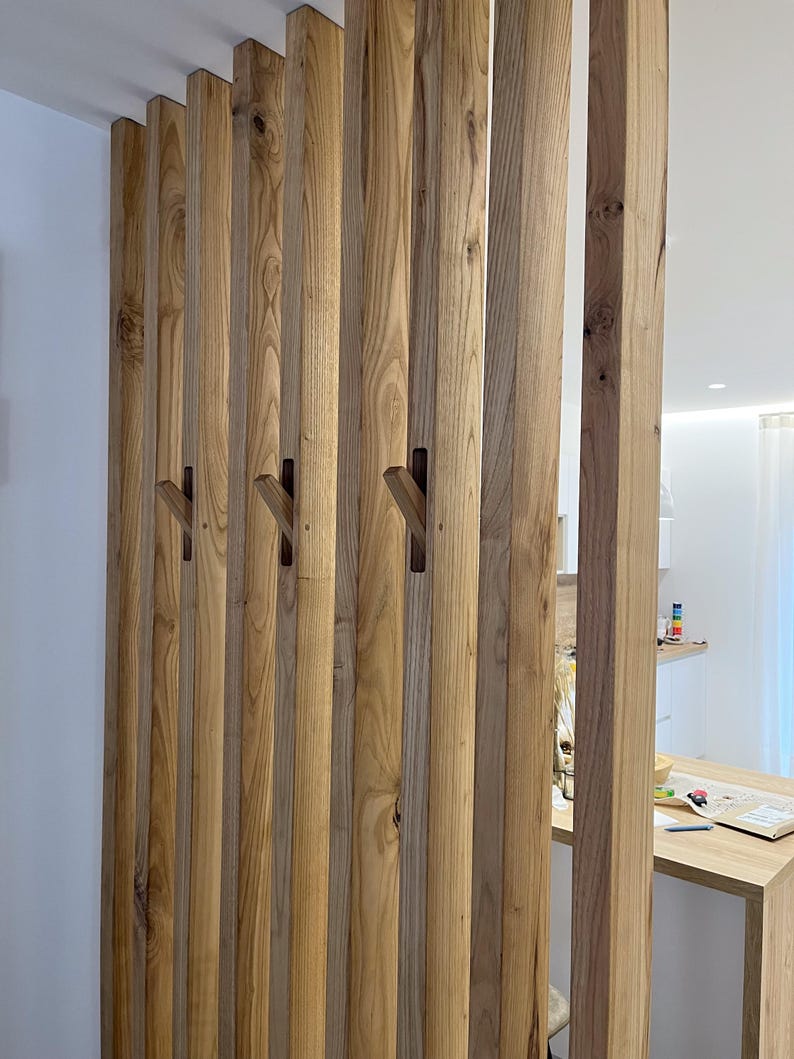 Peut inclure: Un s&eacute;parateur de pi&egrave;ce compos&eacute; de lattes de bois verticales brun clair dispos&eacute;es en parall&egrave;le. Plusieurs de ces lattes pr&eacute;sentent des crochets en bois inclin&eacute;s int&eacute;gr&eacute;s qui d&eacute;passent du centre, offrant une solution de rangement fonctionnelle.