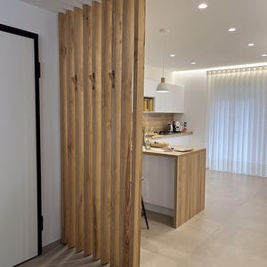 Peut inclure: Un s&eacute;parateur de pi&egrave;ce compos&eacute; de lattes de bois verticales brun clair dispos&eacute;es en parall&egrave;le. La structure int&egrave;gre des crochets en bois fix&eacute;s sur les lattes, cr&eacute;ant une cloison fonctionnelle entre l'espace de vie et la cuisine.