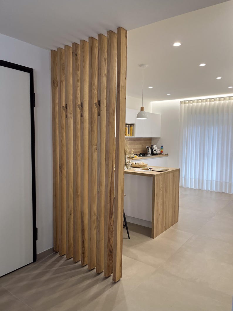 Peut inclure: Un s&eacute;parateur de pi&egrave;ce compos&eacute; de lattes de bois verticales brun clair dispos&eacute;es en parall&egrave;le. La structure int&egrave;gre des crochets en bois fix&eacute;s sur les lattes, cr&eacute;ant une cloison fonctionnelle entre l'espace de vie et la cuisine.