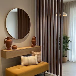 Peut inclure: Un s&eacute;parateur de pi&egrave;ce compos&eacute; de lattes de bois fonc&eacute; verticales, une console flottante en bois clair, un miroir rond &agrave; cadre dor&eacute;, deux vases couleur terre cuite, un banc capitonn&eacute; jaune moutarde et deux coussins d&eacute;coratifs sur le banc.