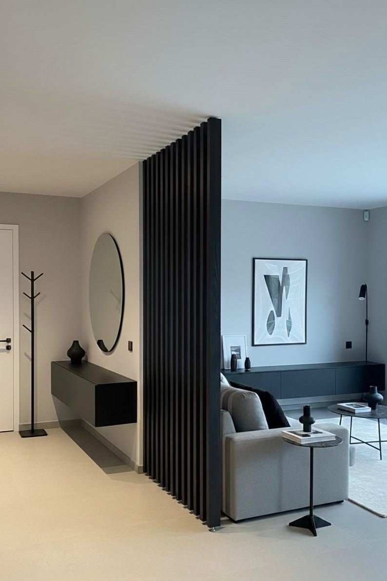 Peut inclure: Un s&eacute;parateur de pi&egrave;ce &agrave; lattes verticales noires se trouve entre un couloir et un salon. Le couloir comprend une console noire suspendue, un miroir rond et un porte-manteau. Le salon contient un canap&eacute; gris, un meuble TV noir et des tables.