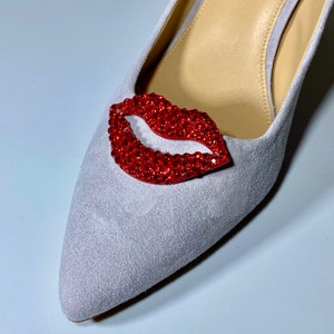 Op de afbeelding: Een lichtgrijze su&egrave;de pump met een rood strass-lipontwerp.
