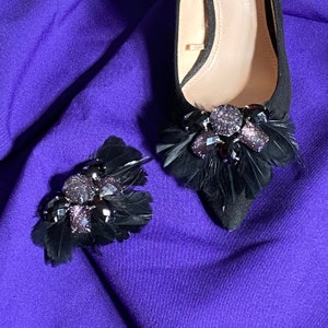 May include: A pair of black suede shoes with black feather and rhinestone embellishments. The shoes are on a purple fabric background.