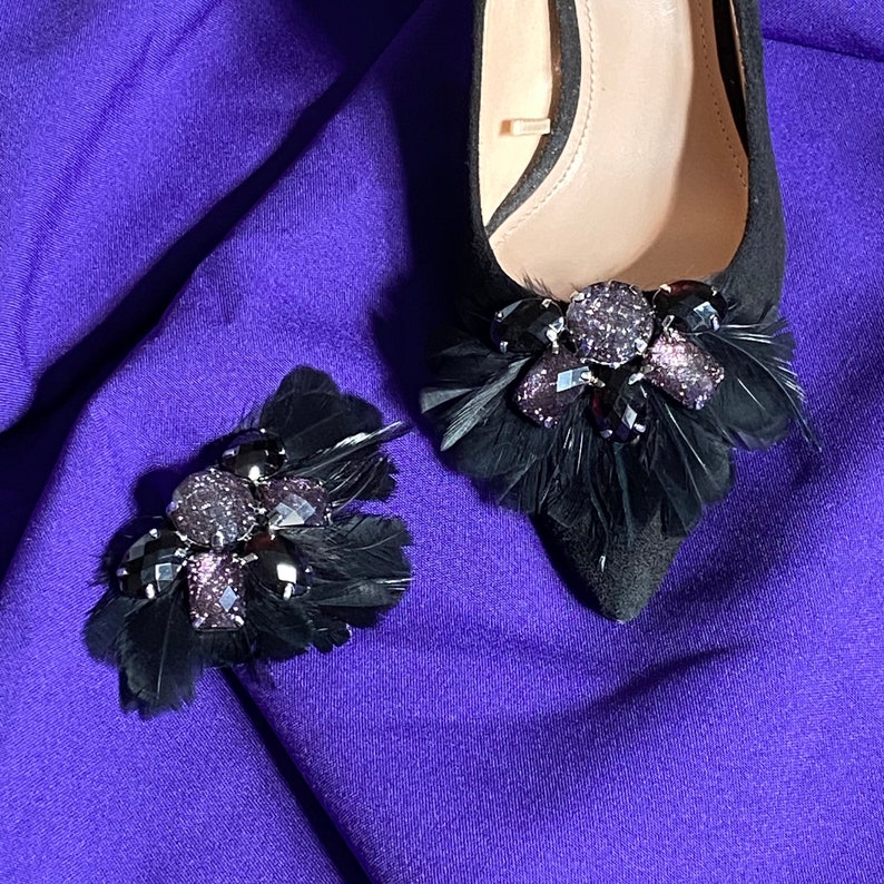 May include: A pair of black suede shoes with black feather and rhinestone embellishments. The shoes are on a purple fabric background.