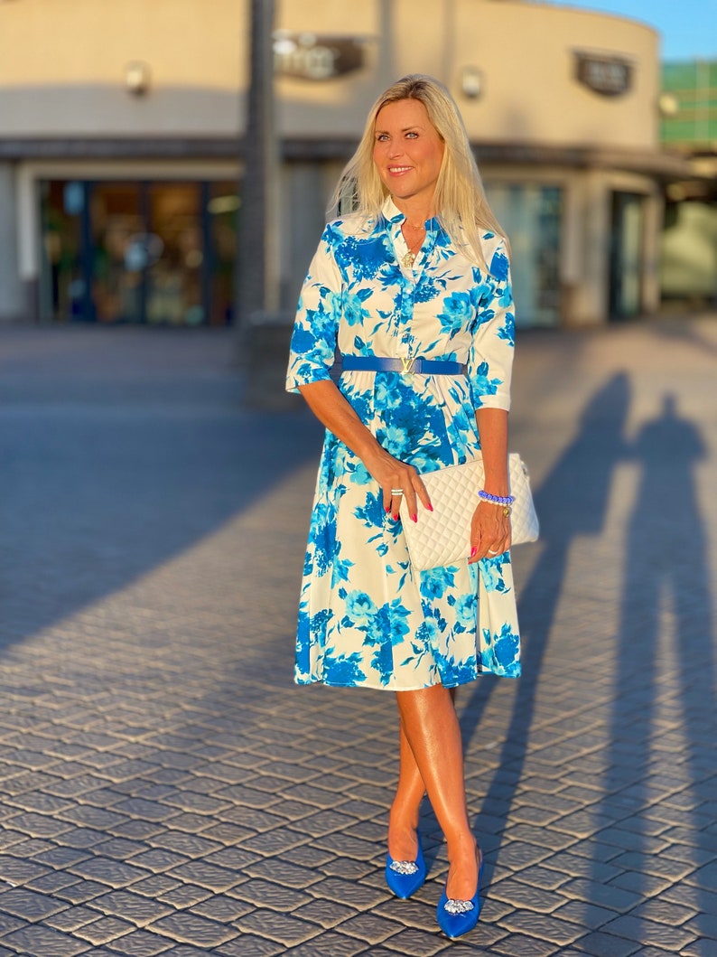 May include: A woman wearing a white dress with a blue floral pattern, a blue belt, and blue shoes. She is holding a white clutch bag.