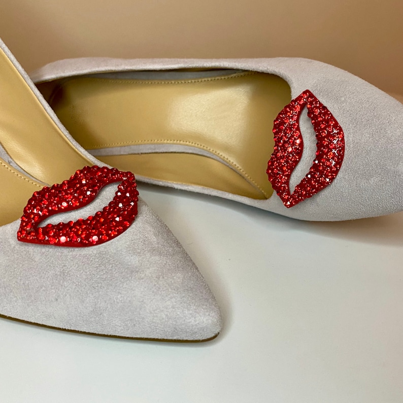 Op de afbeelding: Een paar grijze su&egrave;de schoenen met rode strass-lippen op de tenen.