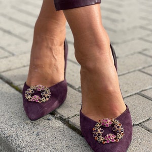 Pode incluir: Um par de sapatos de salto alto de camurça roxo escuro com bico fino e detalhes de joias. Os detalhes são dourados e apresentam uma variedade de gemas coloridas.