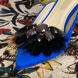 May include: A pair of blue suede heeled mules with a black feather and rhinestone embellishment.
