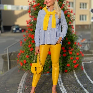 May include: A woman wearing a blue and white striped shirt with a yellow knitted scarf, yellow leather trousers, and yellow shoes. She is carrying a yellow handbag with a gold chain strap.