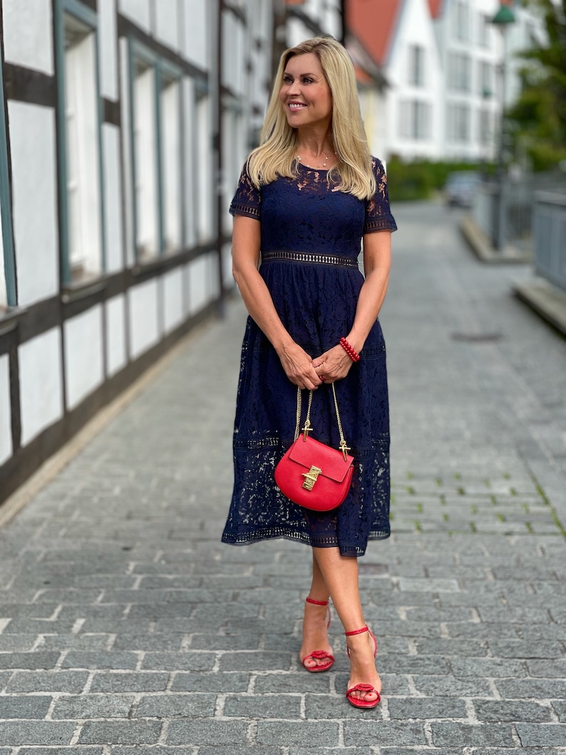 Op de afbeelding: Een vrouw in een donkerblauwe kanten jurk met een rode handtas en rode hakken. Ze staat op een straat met keien.
