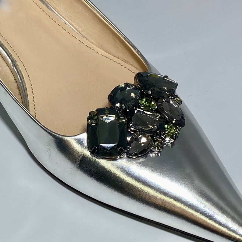 May include: Silver leather pointed toe flat shoes with a cluster of dark grey and green rhinestones on the toe.