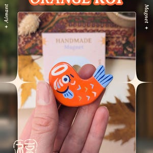 May include: An orange koi fish magnet with blue and white details. The magnet is part of the "Koi / Totoro Collection" and is handmade. The text "Orange Koi" is displayed above the magnet. The artist's name, "Art by Asudraws", is at the bottom.