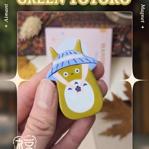 May include: A Green Totoro magnet from the Koi / Totoro Collection. The magnet features a yellow Totoro character with a blue and white hat. The text "GREEN TOTORO" is displayed above the character. The art is by Asudraws.