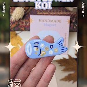 May include: A light blue koi fish magnet with a floral design. The magnet has a white eye and is decorated with gold and white flowers. Text on the image reads "BLUE FLOWER KOI" and "HANDMADE Magnet". The art is by Asudraws.