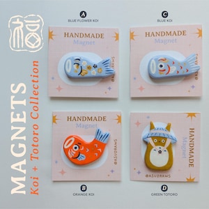 Peut inclure: Collection d'aimants faits à la main en argile séchée à l'air, représentant des carpes koï et un personnage Totoro vert. Les aimants sont présentés sur des cartes roses et comprennent un koï bleu fleuri, un koï bleu, un koï orange et un Totoro vert. Texte : "MAGNETS Koi + Totoro Collection".