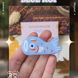 May include: A light blue koi fish magnet with orange and white details. The magnet is part of the "Blue Koi" collection and is handmade. The image also includes the text "Aimant" and "Magnet".