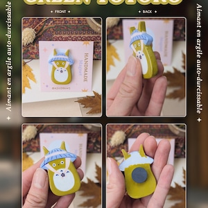 May include: A handmade green Totoro magnet from the Koi / Totoro Collection. The magnet features a yellow Totoro character with a blue hat and white belly. The text "GREEN TOTORO" is displayed above the magnet. The text "Art by Asudraws" is displayed below the magnet.