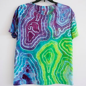 Puede incluir: Una camiseta tie-dye con un patr&oacute;n de remolino de morado, azul, verde y blanco.