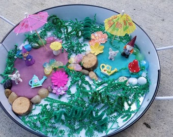 May include: A white tray with two sections of play dough, one pink and one blue, decorated with miniature figurines, flowers, butterflies, and umbrellas. The tray is filled with green grass-like material.