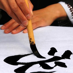 May include: A person's hand holding a traditional Japanese calligraphy brush, writing black ink characters on white paper. The brush is made of bamboo and has long black bristles. The characters are partially formed and appear to be the Japanese word "fun".