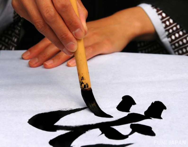 May include: A person's hand holding a traditional Japanese calligraphy brush, writing black ink characters on white paper. The brush is made of bamboo and has long black bristles. The characters are partially formed and appear to be the Japanese word "fun".