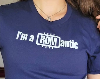 May include: A woman wearing a navy blue t-shirt with the text "I'm a ROM antic" and a white graphic of a computer chip. She is also wearing denim shorts and clear glasses.