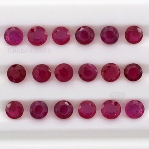 May include: Sixteen round, faceted rubies in varying shades of red and pink are arranged in rows. The gemstones are displayed on a white surface. The text "KHADKA GEMS" is visible in the image.