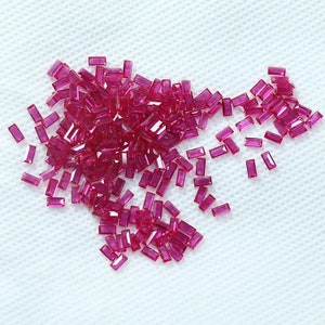 May include: A large quantity of small, rectangular, red ruby gemstones. The gemstones are scattered on a white surface.