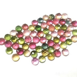 May include: A collection of faceted gemstones in various colors, including pink, green, yellow, and clear. The gemstones are round and appear to be tourmaline, scattered on a white surface. The gems have a polished, reflective surface.