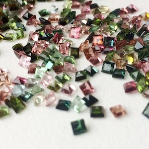 May include: A scattering of small, square-cut gemstones in shades of pink, green, and brown. The gems are spread across a white surface, creating a colourful and textured display. These gems are likely for jewellery making.