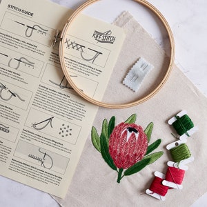 May include: A close-up of a embroidery kit with a wooden hoop, a piece of white fabric, and a variety of embroidery floss. The fabric has a red and green embroidered flower design. The kit includes instructions for different embroidery stitches, including "Couching", "Seed Stitch", and "Buttonhole/Blanket Stitch".