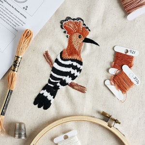 May include: A close-up of a hoop embroidery project featuring a colourful bird with black and white stripes. The embroidery is done on a beige fabric and includes a wooden embroidery hoop, a thimble, and spools of embroidery floss in white, black, and orange.