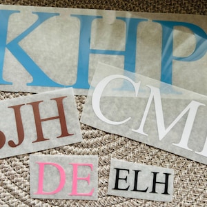 May include: Five sheets of white paper with different letters cut out of them. The letters are in different colours, including blue, brown, white, pink, and black. The letters spell out the names "KHP", "CMR", "SJH", "DE", and "ELH".