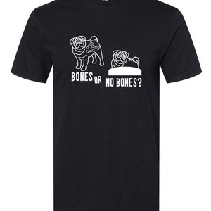 Op de afbeelding: Zwart T-shirt met een witte afbeelding van twee mopshonden. Eén mopshond heeft botten achter zich en de andere niet. De tekst "Bones or No Bones?" staat onder de mopshonden.