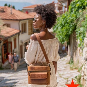 May include: A brown leather crossbody bag with a top handle and front pocket. The bag has a shoulder strap and a zipper closure. The bag is worn by a person in a striped off-the-shoulder top and shorts.