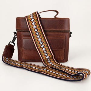Könnte beinhalten: Eine strukturierte braune Ledertasche mit einem Tragegriff und einer Vordertasche. Die Tasche verfügt über einen abnehmbaren, breiten, gewebten Schulterriemen mit einem geometrischen Muster in Orange, Grün, Weiß und Dunkelblau, befestigt mit Metallclips.