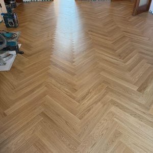 Puede incluir: Suelo de parquet de madera clara con patrón de espiga. Las tablas están dispuestas en un diseño en forma de V repetida, creando una textura visualmente atractiva. La veta de la madera es visible, añadiendo calidez. El suelo está instalado en una habitación luminosa.