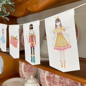 Puede incluir: Una pancarta con temática de Cascanueces con ilustraciones de acuarela de una bailarina, un tambor, un soldado cascanueces y un hada de azúcar. La pancarta está enhebrada en una cuerda blanca y está colgada de un estante de madera.