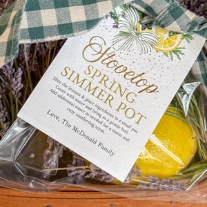 Puede incluir: Una cesta de regalo con un lazo a cuadros verdes y blancos, que contiene lavanda, un limón y una etiqueta que dice "Stovetop Spring Simmer Pot". La cesta está hecha de material marrón tejido.