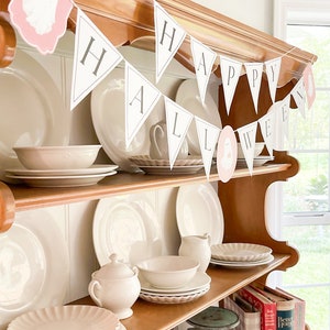 May include: A wooden hutch with white dishes and a banner that reads "Happy Halloween". The banner is made of white paper with black letters and features pink ghost cutouts.
