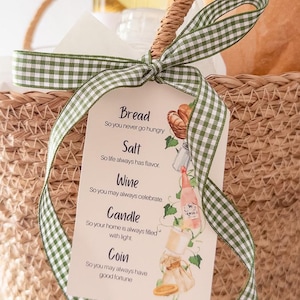 Puede incluir: Una cesta de mimbre con una cinta de cuadros verdes y blancos atada con un lazo. Una etiqueta con ilustraciones de pan, sal, vino, una vela y una moneda está adjunta. La etiqueta dice "Bread, Salt, Wine, Candle, Coin."