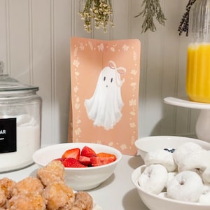 May include: A white table with a variety of food items, including donuts, strawberries, and fried dough balls. A sign with a cartoon ghost with a pink bow is on the table.
